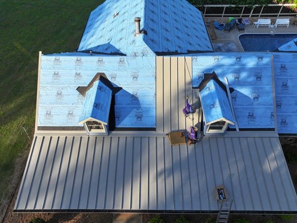 An aerial view of a house roof under construction with silver metal panels and blue underlayment, plus two workers.