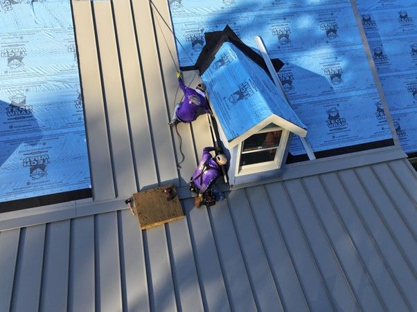 Two workers in purple safety gear install metal roofing panels around a dormer window on a residential roof.