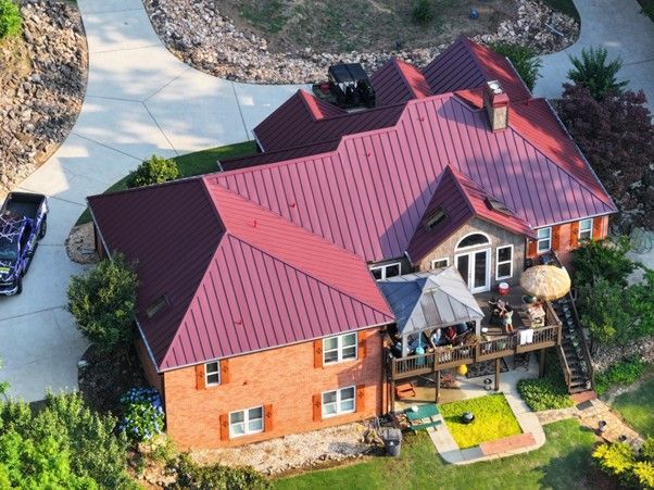 An aerial view of a two-story brick house with a dark red metal roof, surrounded by a paved driveway and green landscaping.
