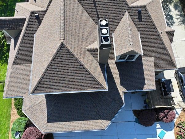 Aerial view of a brown shingled residential roof with multiple peaks, dormers, a chimney, and a small patio below.