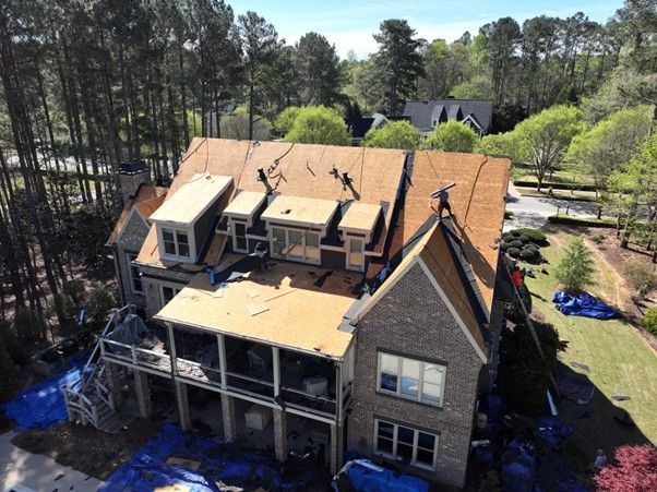 An aerial view of a large residential home under renovation, showing workers on a partially shingled roof.