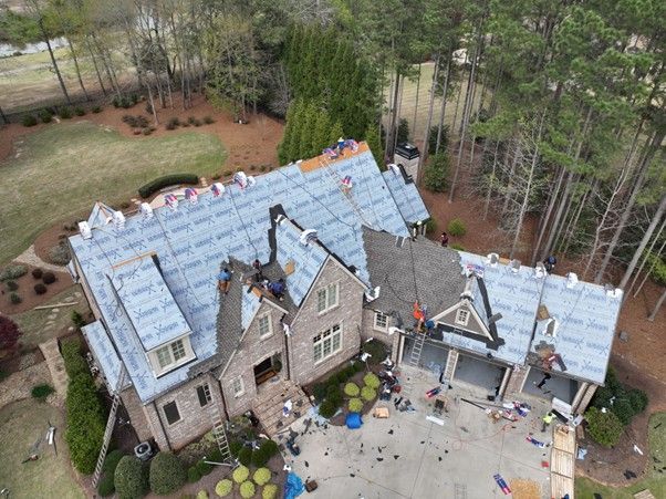 An aerial view of a house under construction with workers installing new roofing material on the bare wooden structure.