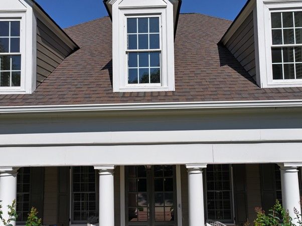 A house exterior featuring a covered porch with white columns, brown shingled roof, and three dormer windows.