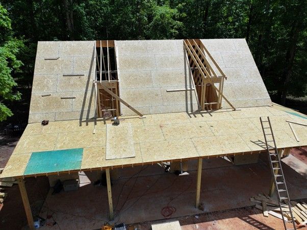 An elevated view of a wooden roof under construction with two open dormer frames and a ladder leaning against the side.