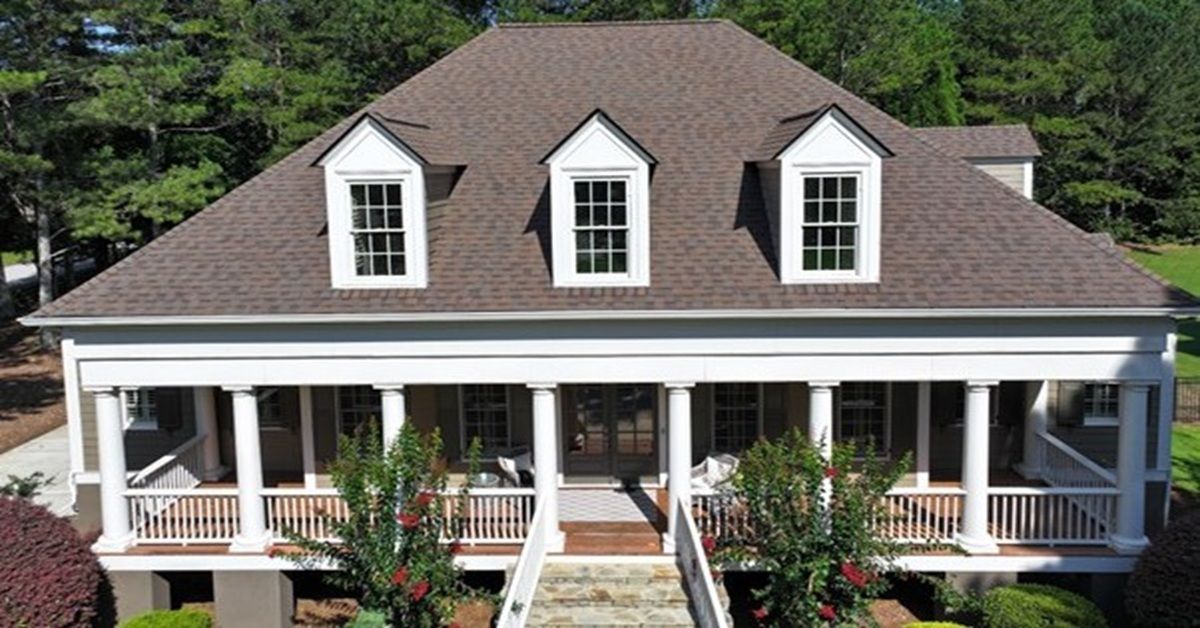 A two-story house with a white wraparound porch, columns, and three dormer windows on a brown shingled roof.