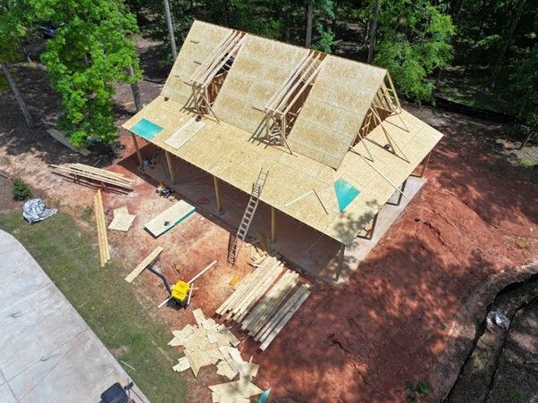 An aerial view of a wooden structure under construction in a wooded area, showing rafters and partially sheathed roof.