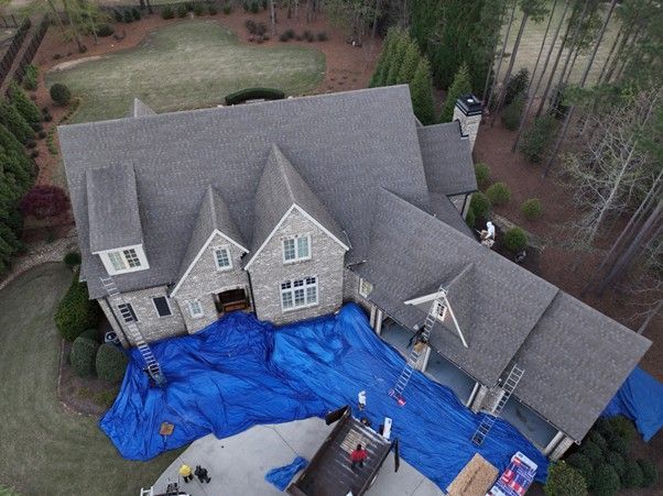 Aerial view of a home with a gray shingled roof under construction, protected by large blue tarps over the driveway.