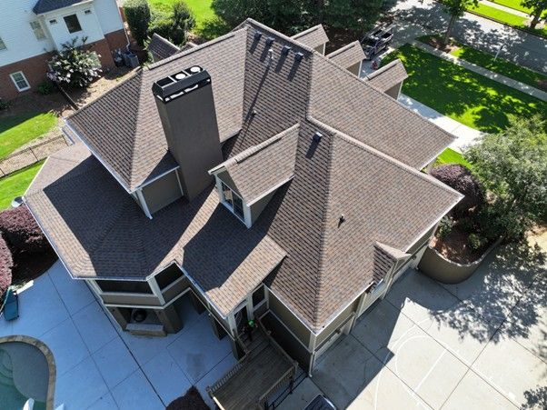 An elevated, high-angle aerial view of a dark brown residential shingle roof with a chimney and a small dormer window.