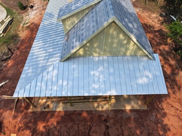 An aerial view of a metal-roofed house under construction, with a gabled dormer and reddish-brown dirt ground.
