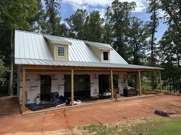 A partially constructed house frame with a metal roof and two dormers, surrounded by trees on a dirt lot.