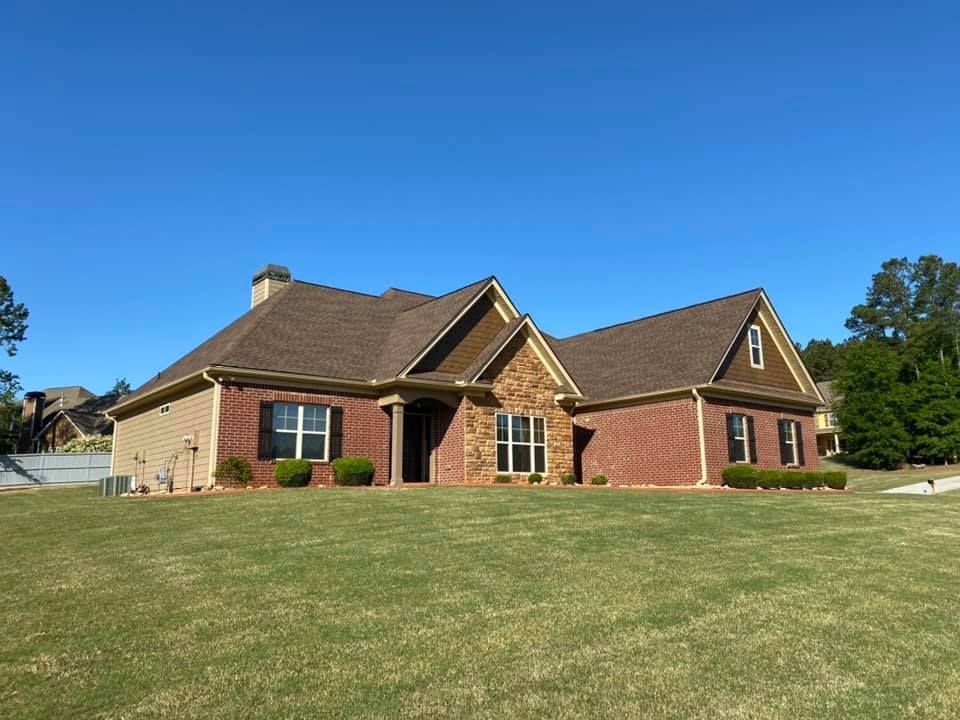 Brick and tan home with brown roof, stone accents, and green lawn under blue sky.
