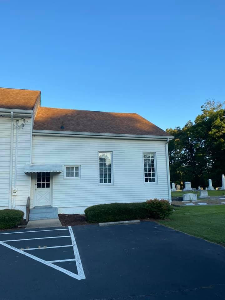 White church building with brown roof, door, and windows next to a cemetery and blue sky.