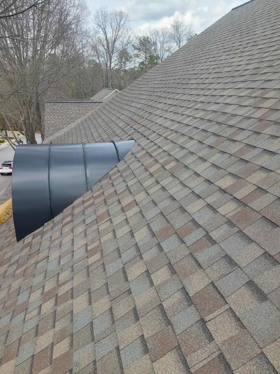 Brown and grey shingled roof with a large black exhaust pipe. Trees in the background.
