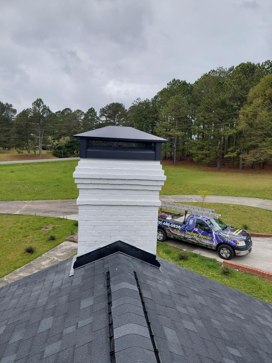 View of a gray shingled roof with a white chimney, topped with a black cap, and a truck in the background.