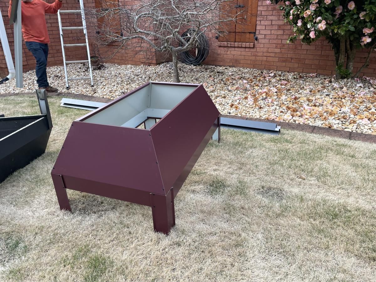 A maroon metal planter box on grass, with a person and ladder in the background.