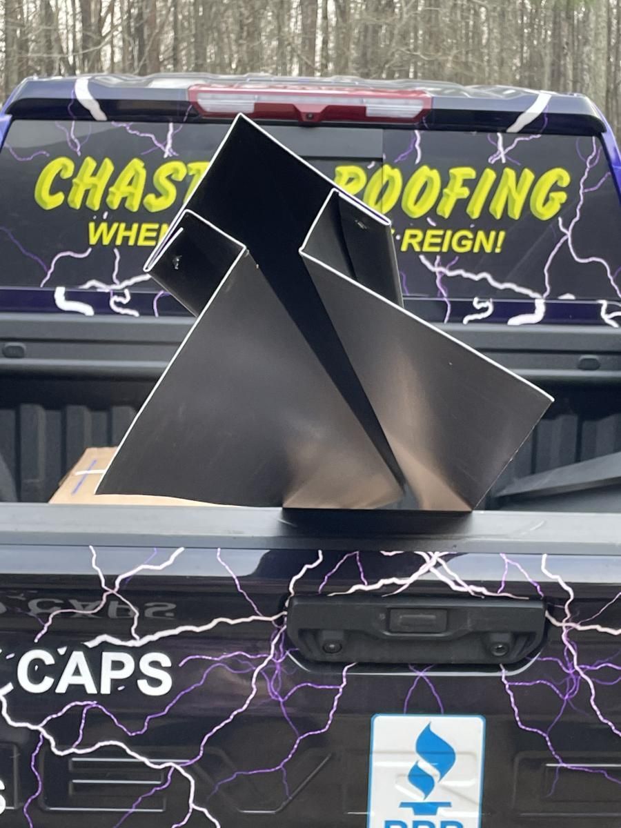 A metallic, angular roof flashing sits in the bed of a truck with roofing company logos and a lightning bolt design.