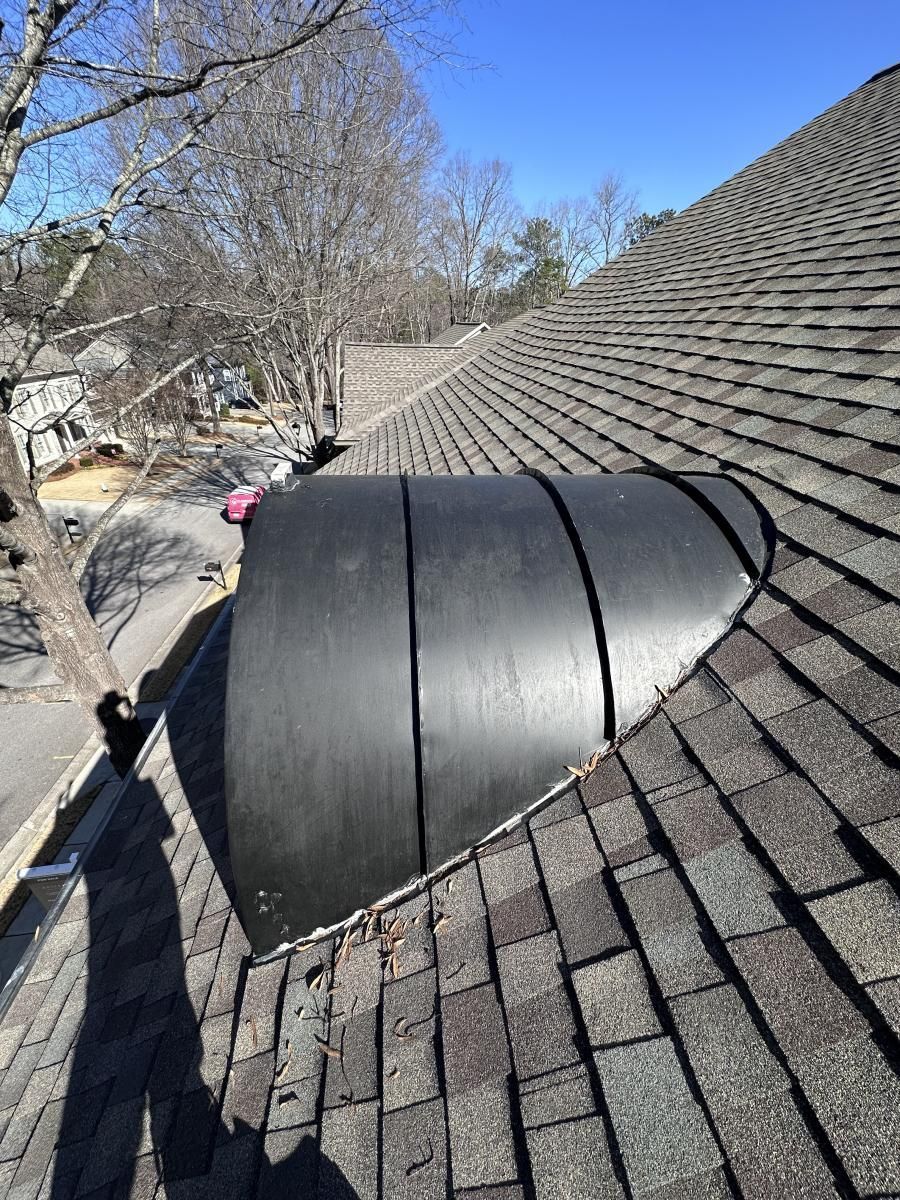 Black cylindrical structure on a gray shingled roof with bare trees and a street in the background.