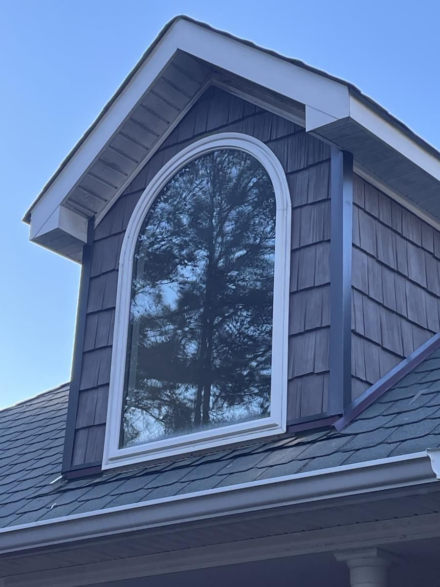 Gabled dormer with arched window, dark siding, white trim, blue sky, and a tree reflected in the glass.