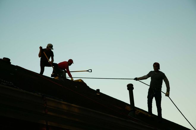 Silhouetted roofers work on a roof against a clear sky, using tools to make repairs.