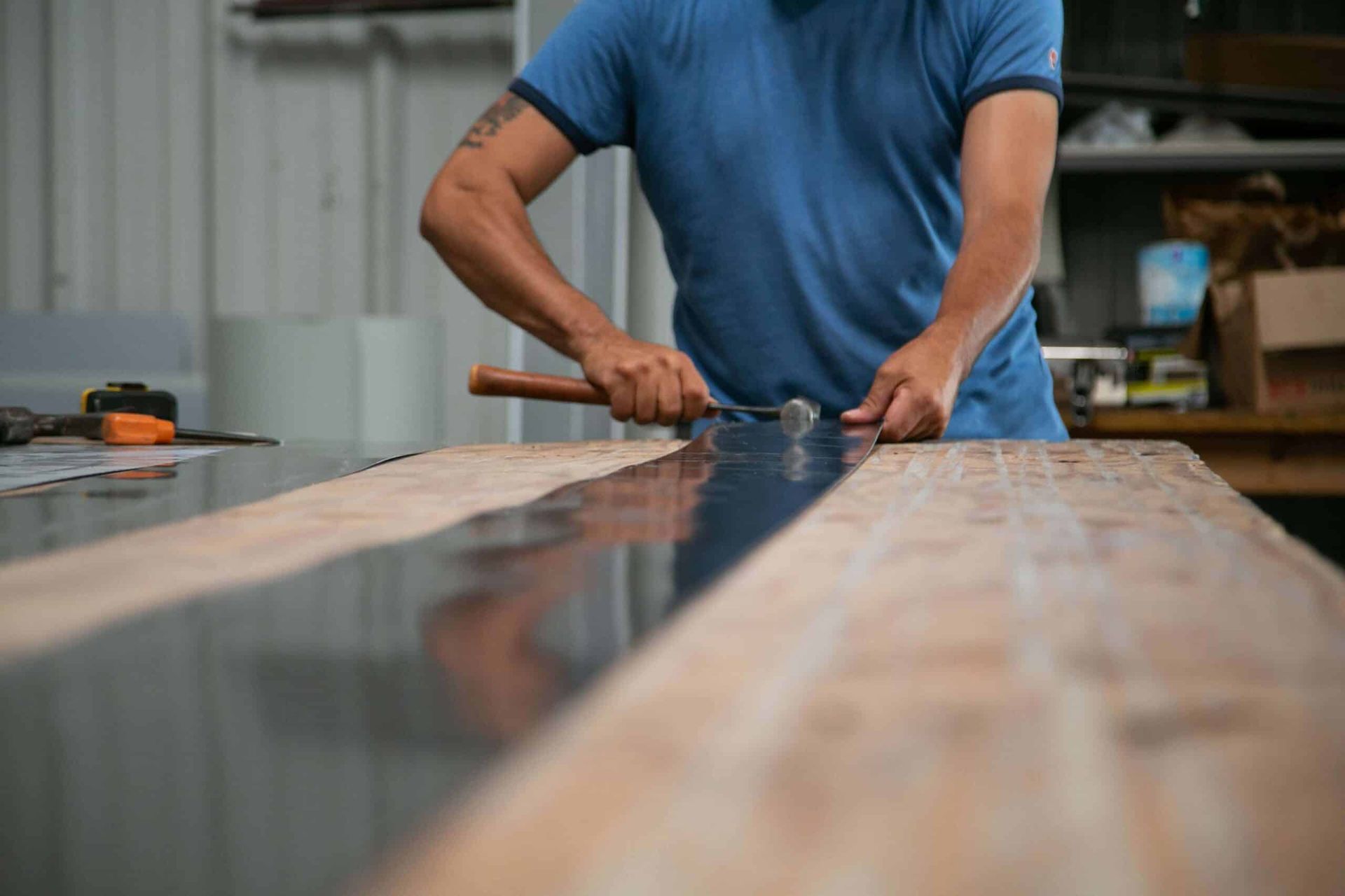 Man in blue shirt using a hammer on a wood surface in a workshop.