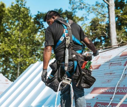 Roofer wearing safety harness on a roof, holding white material. Trees and blue sky in background.