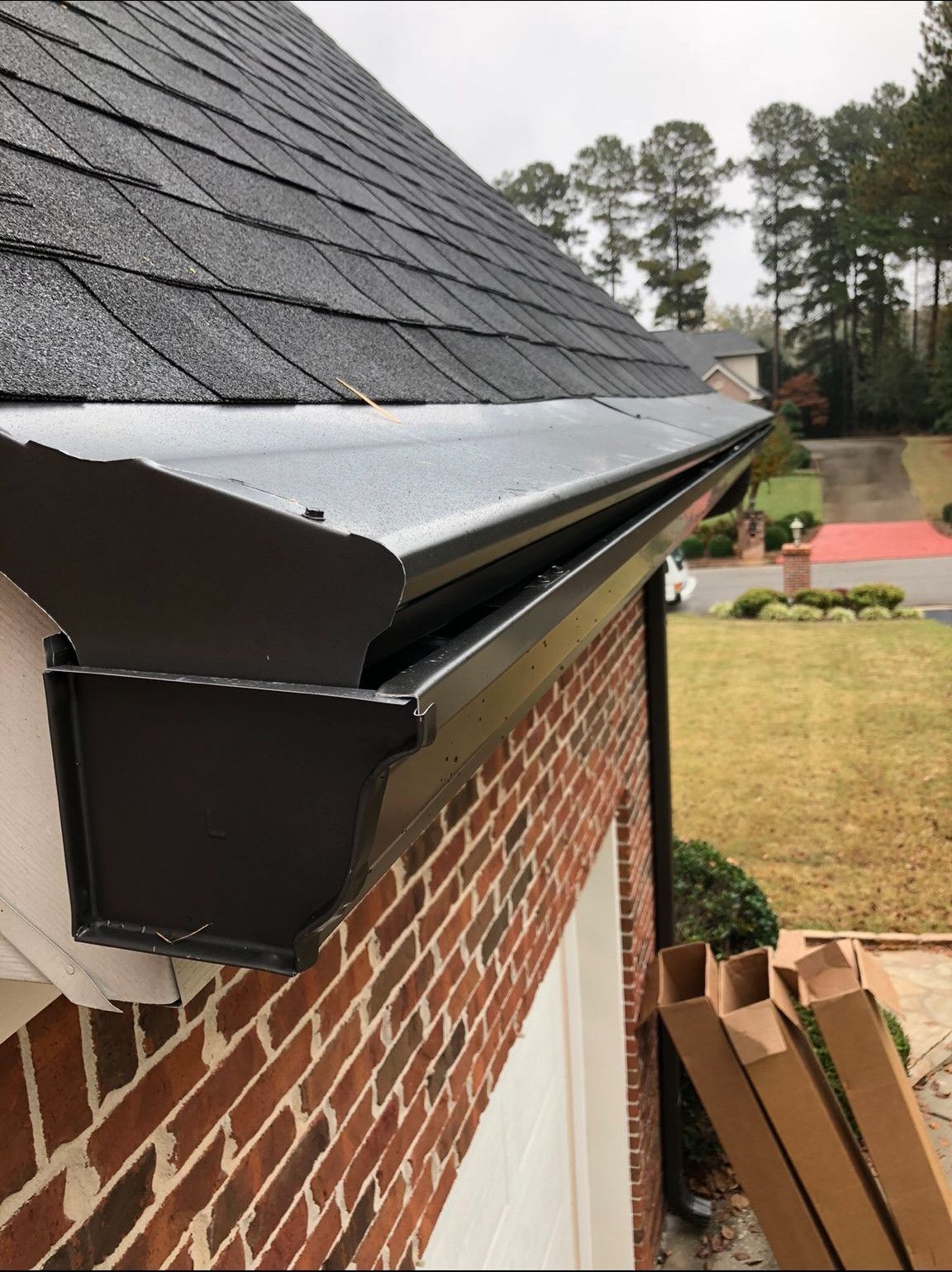 Black gutter along a brick house, beneath a dark shingled roof. Brown cardboard boxes are on the ground.