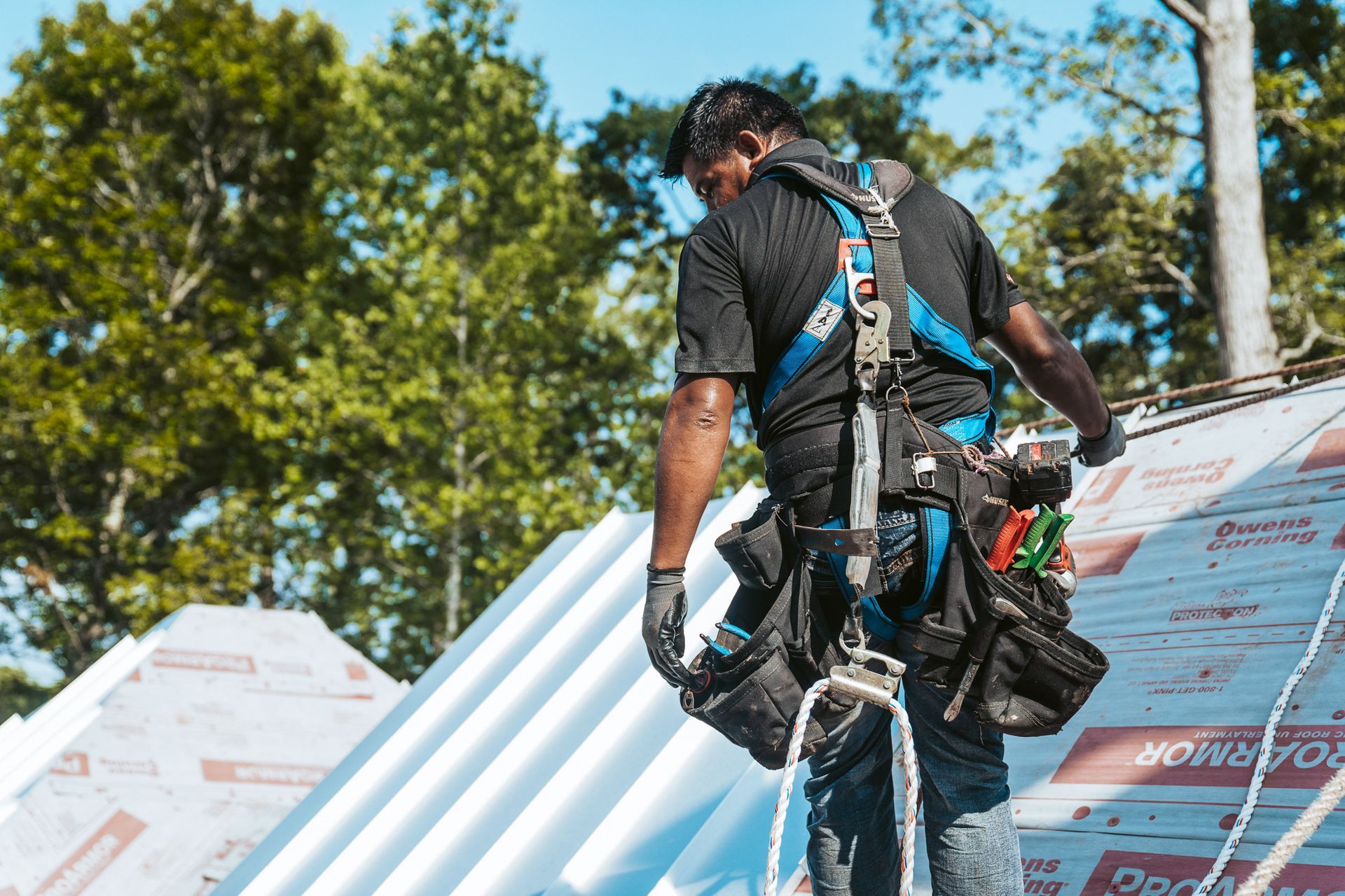 Roofer wearing safety harness working on a roof; blue harness, gray roof, tools.