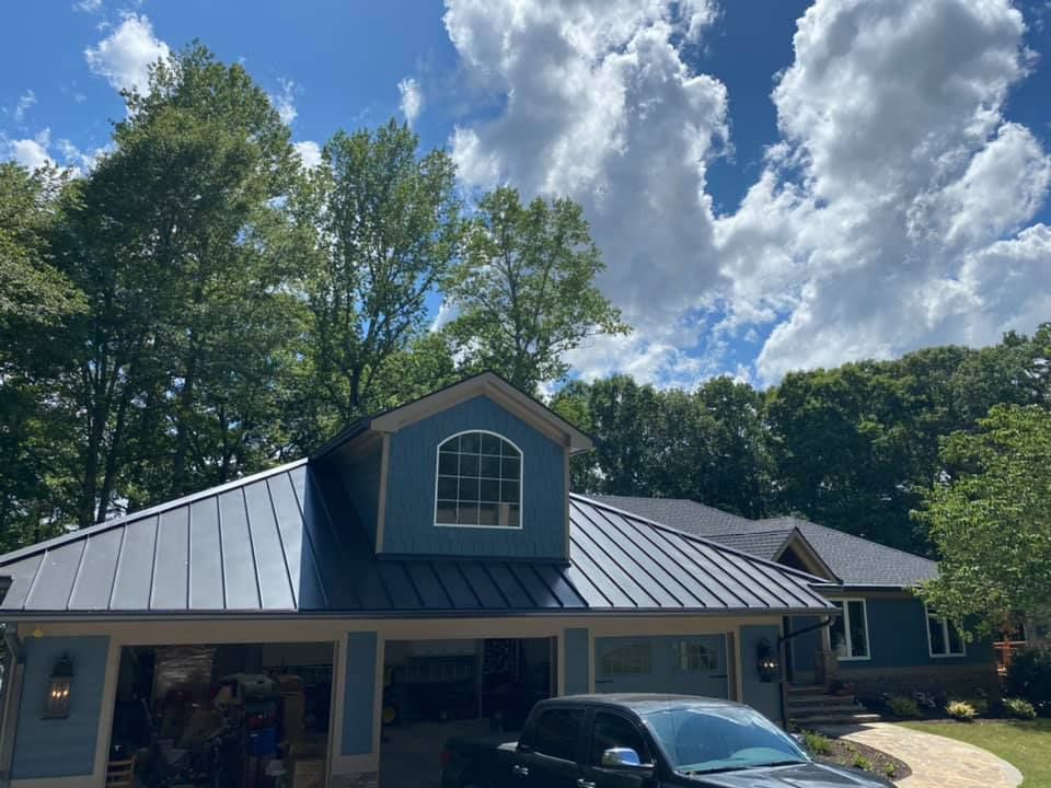 Blue house with a metal roof, garage, and a truck parked in front; blue sky and trees in background.