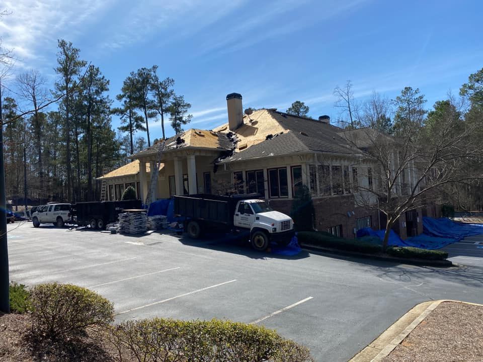 Roof being repaired on a building; dump truck and materials on site. Blue sky, trees.