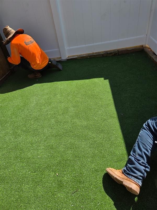 Person installing artificial turf in a small, fenced-in outdoor area with a second person seated nearby.