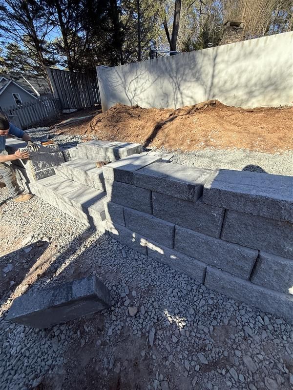Stone retaining wall construction in progress; gravel base, stacked gray blocks, person working.