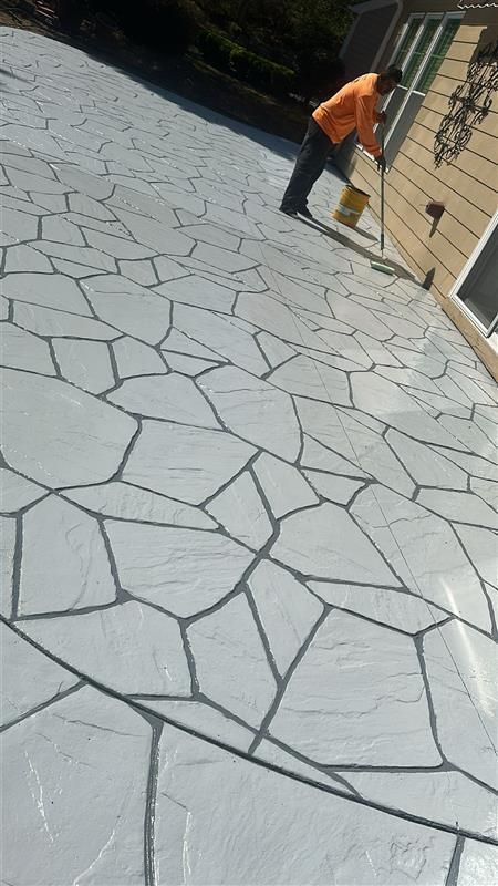 Person applying sealant to a patterned concrete patio. Gray and black design, light yellow building.