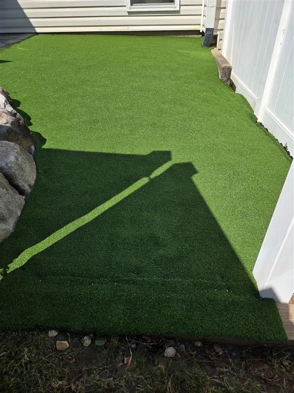 Green artificial turf installed in a small backyard area between a house wall and a white fence.