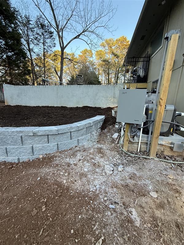 Retaining wall made of gray blocks, next to a building and dirt yard with exposed wiring.