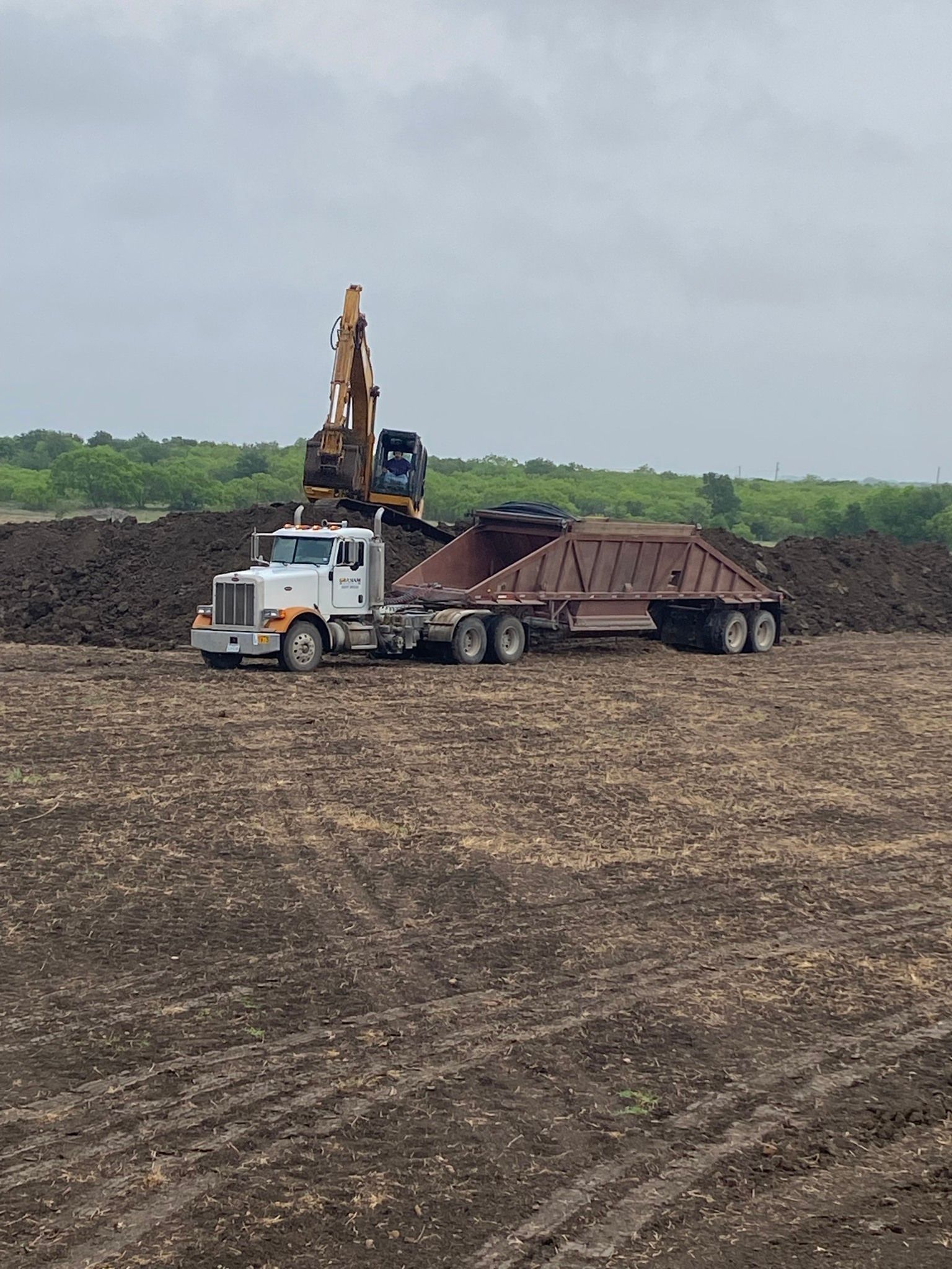 A dump truck is driving through a dirt field with a bulldozer in the background.