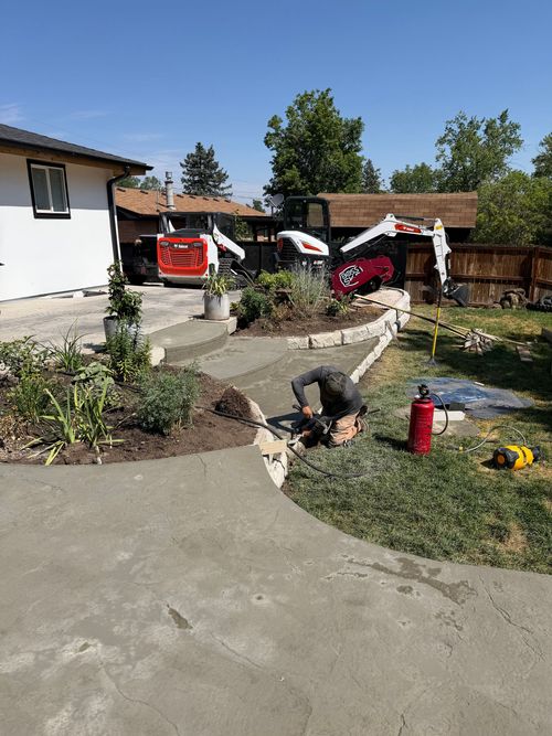 Two workers in a yard repairing a sprinkler near a red fire extinguisher and hoses by a house