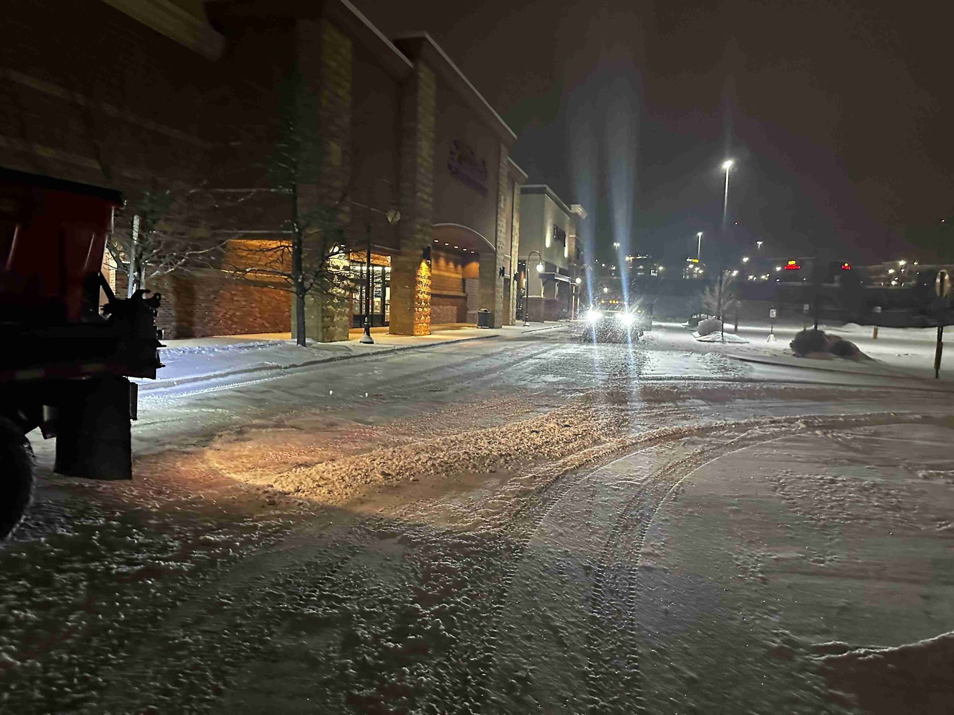 Snow-covered street at night beside a lit brick building, with falling snow and car headlights in the distance