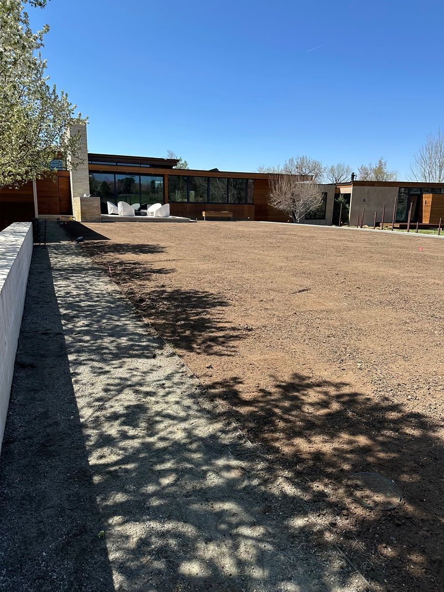 Wide outdoor courtyard with gravel ground, shaded trees, and a low modern building under a clear blue sky