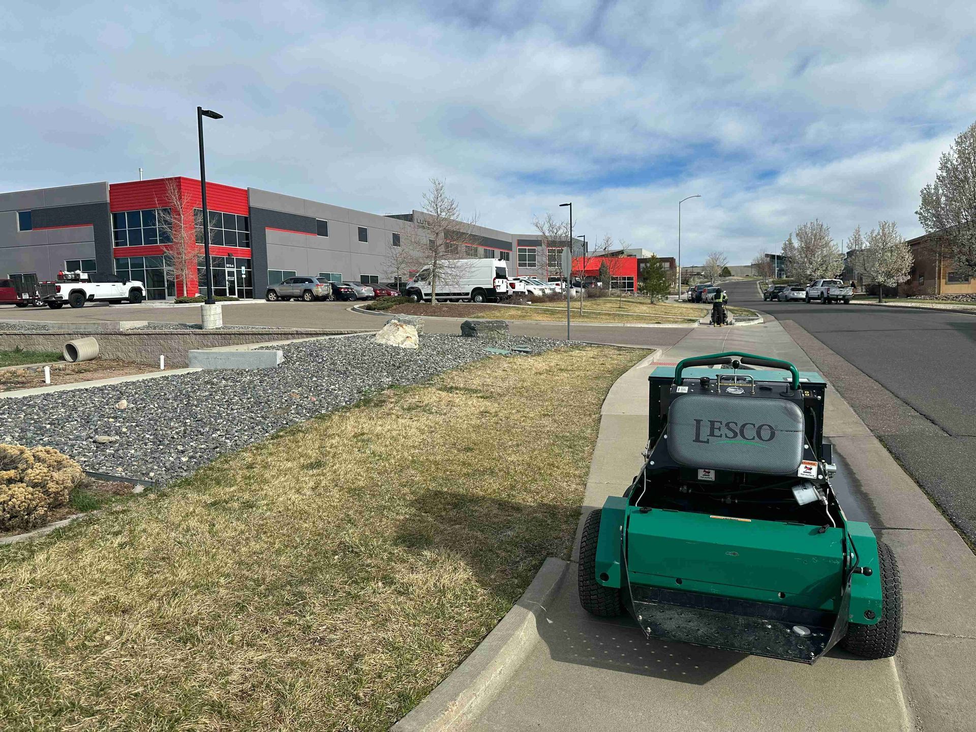 Green lawn mower on a sidewalk beside a road, with a red-and-gray building in the background