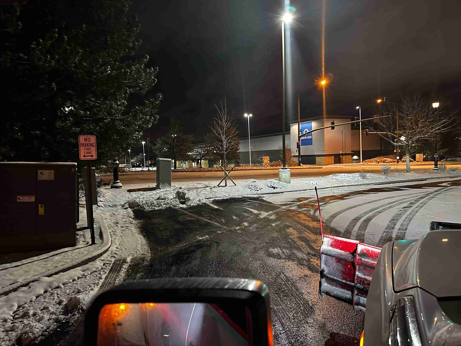 Snowy parking lot at night with streetlights, parked cars, and a red-and-white barrier near the curb