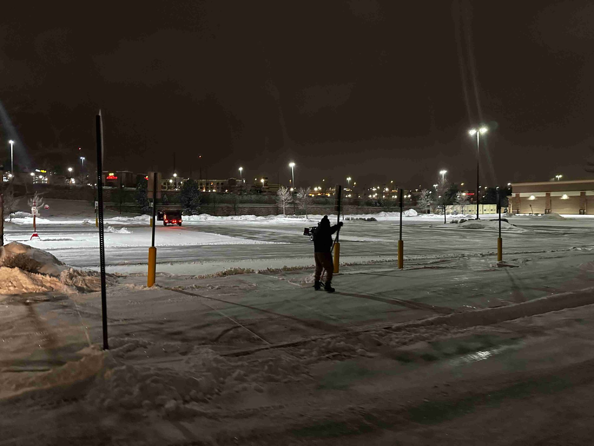 Snowy parking lot at night with one person standing under streetlights near parked cars.