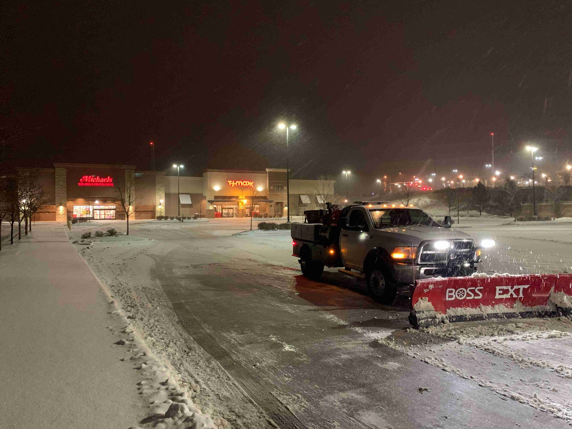 Snowy parking lot at night with a plow truck clearing snow near lit storefronts
