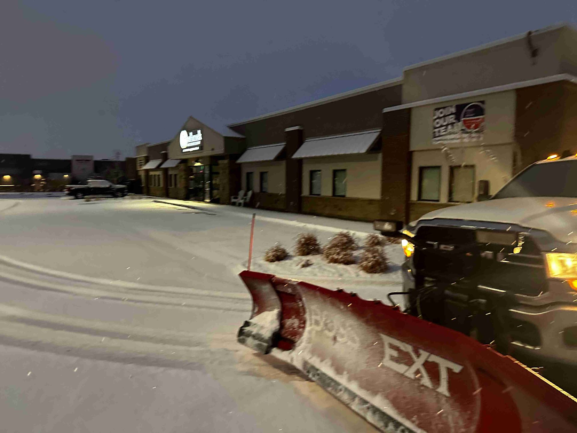 Snow-covered shopping center at night with a red snowplow blade in the foreground and lit storefronts beyond