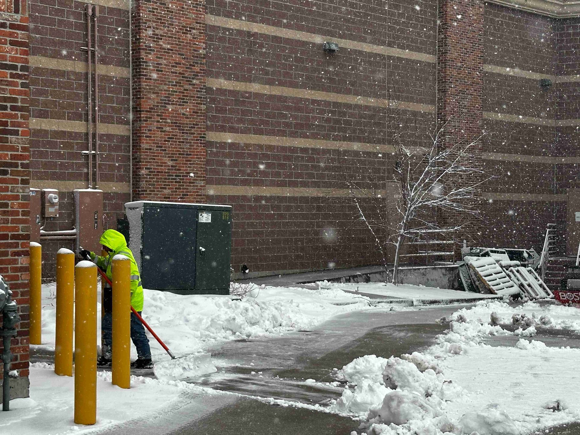 Snowy alley beside a brick building with yellow bollards and a utility box