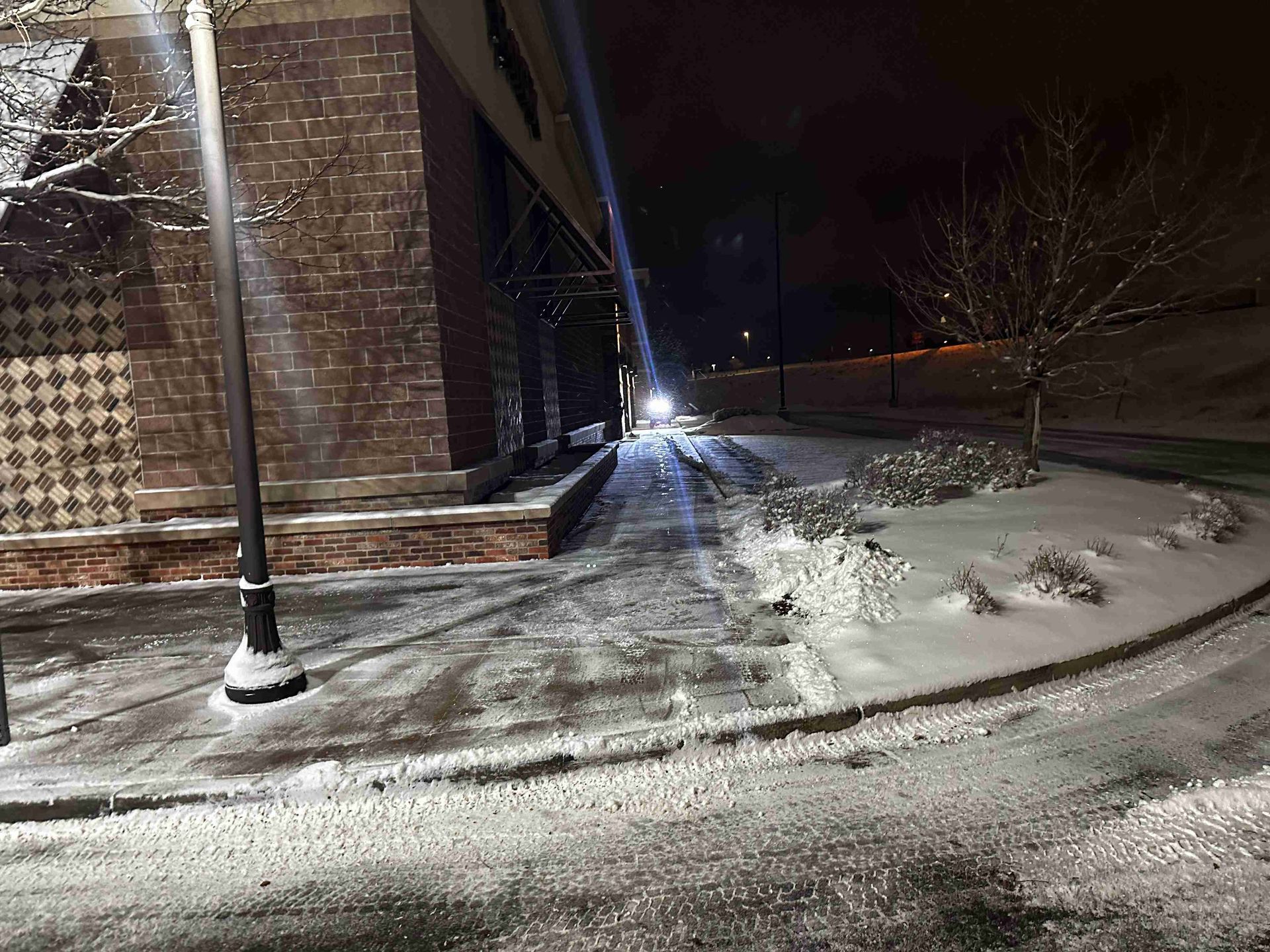 Snowy night beside brick building and streetlamp, with a lit path along the corner.