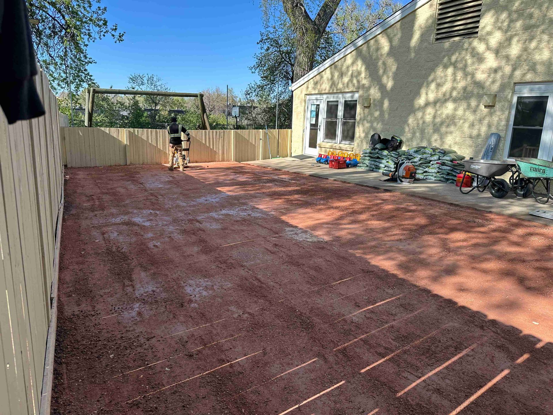 Backyard with red dirt ground, a person near the fence, and a house with chairs and yard items by the wall