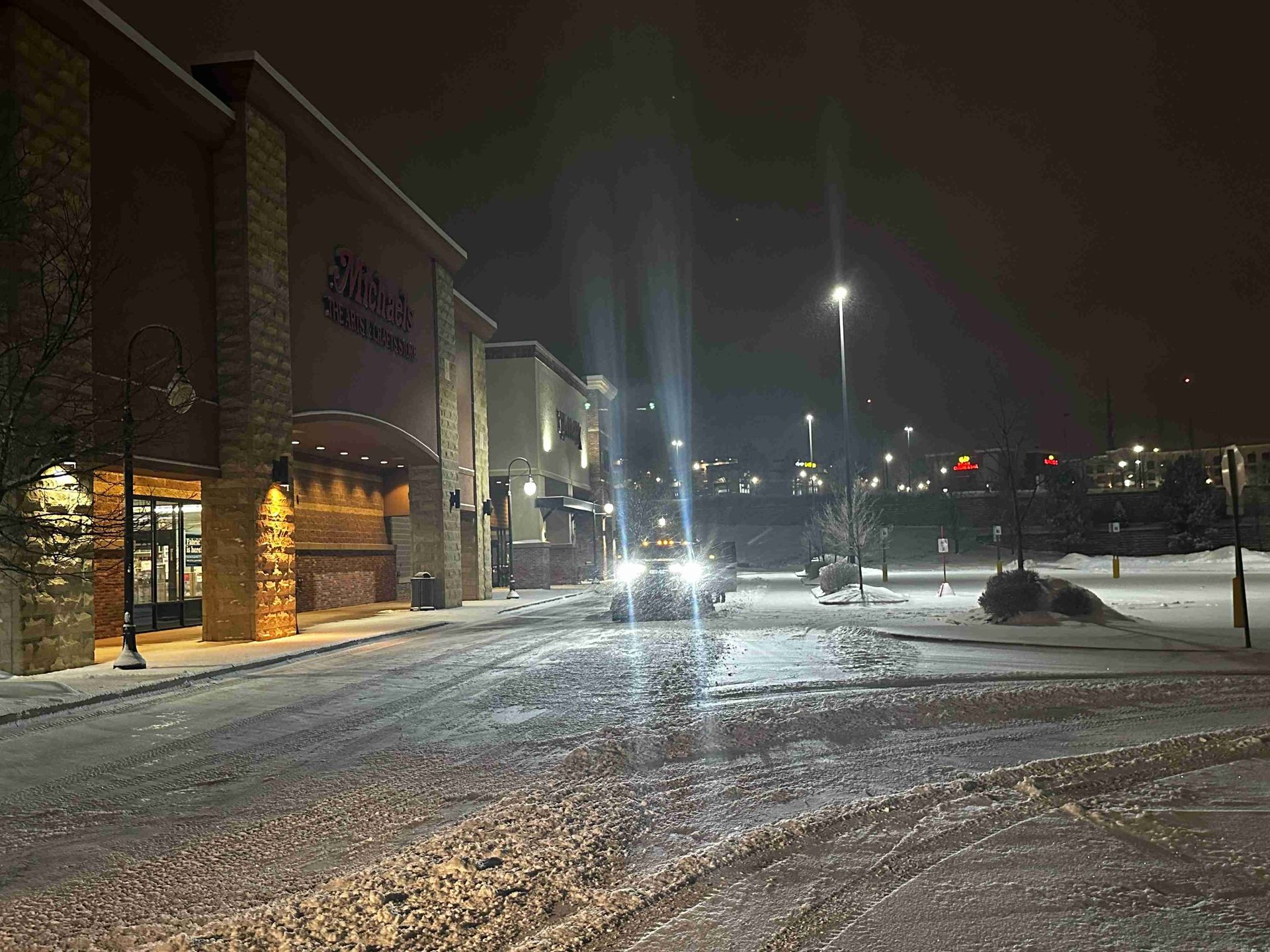 Snowy parking lot at night with a lit building entrance and streetlights reflecting on wet pavement