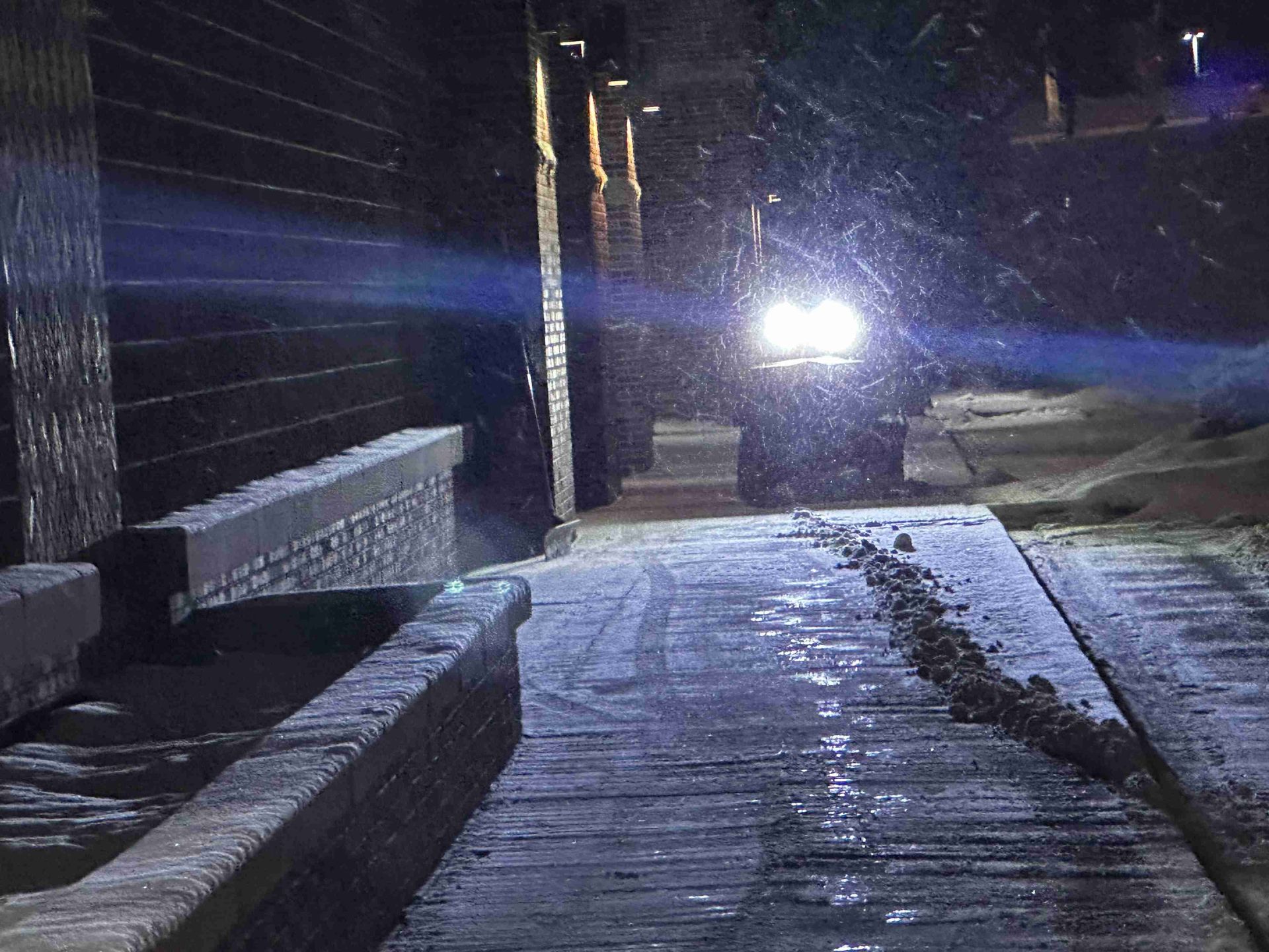 Snow-covered walkway at night with bright headlights shining toward the camera and a bench on the left.