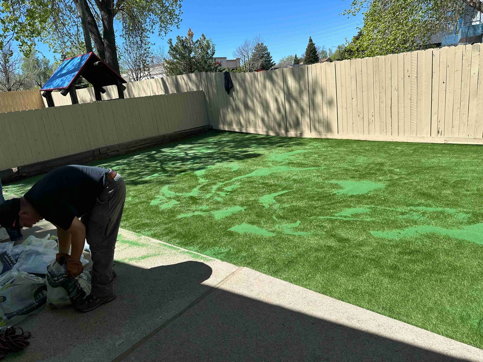 Worker spreading green turf in a fenced backyard under a clear blue sky