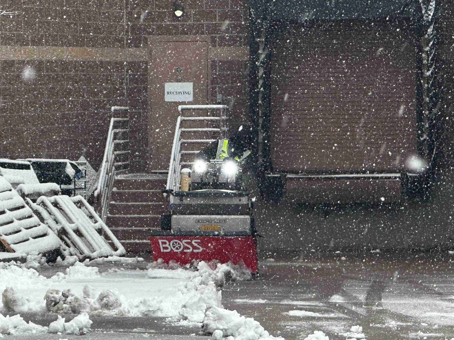 Snowplow clearing heavy snow in a parking lot near brick steps and a building entrance